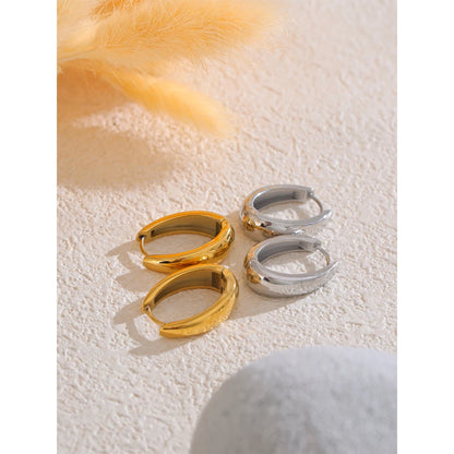 Two pairs of chunky huggie hoops, one gold-tone and one silver-tone, on a beige surface with dried grass