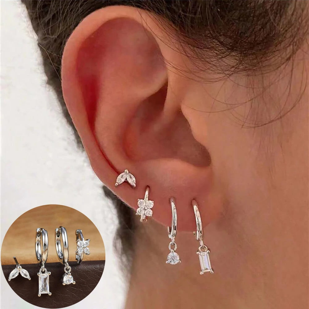 Ear wearing a silver-tone stack of clear-stone leaf stud, flower huggie and two drop hoops, with inset of the set